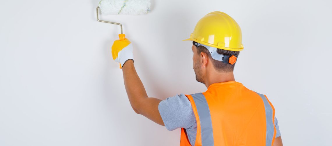 Male builder in uniform, helmet, gloves painting wall with roller , back view.